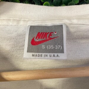 Pu&ograve; includere: Primo piano di una maglietta bianca con etichetta Nike. L'etichetta presenta il logo Nike rosso e la scritta "S (35-37)" e "MADE IN U.S.A.". La maglietta &egrave; appesa a un'asta di legno.