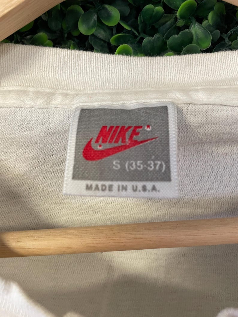 Pu&ograve; includere: Primo piano di una maglietta bianca con etichetta Nike. L'etichetta presenta il logo Nike rosso e la scritta "S (35-37)" e "MADE IN U.S.A.". La maglietta &egrave; appesa a un'asta di legno.