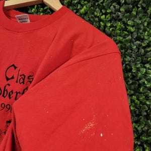 May include: Red t-shirt with black text that reads "Clas Ober" and the number "99". The shirt is hanging on a wooden hanger against a green background. The shirt has some white spots on the sleeve.