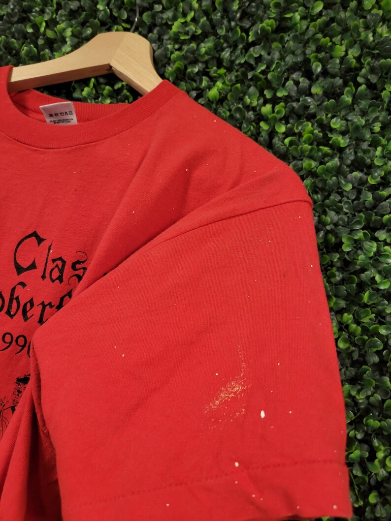May include: Red t-shirt with black text that reads "Clas Ober" and the number "99". The shirt is hanging on a wooden hanger against a green background. The shirt has some white spots on the sleeve.