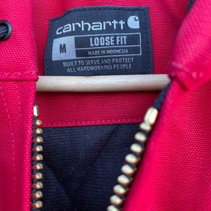 Pode incluir: Close-up de uma jaqueta Carhartt vermelha com forro interno preto e z&iacute;per dourado. A etiqueta da jaqueta diz "carhartt", "LOOSE FIT" e "MADE IN INDONESIA". A etiqueta tamb&eacute;m diz "BUILT TO SERVE AND PROTECT ALL HARDWORKING PEOPLE".