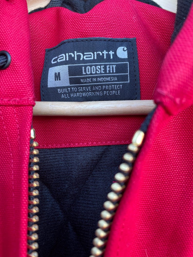 Pode incluir: Close-up de uma jaqueta Carhartt vermelha com forro interno preto e z&iacute;per dourado. A etiqueta da jaqueta diz "carhartt", "LOOSE FIT" e "MADE IN INDONESIA". A etiqueta tamb&eacute;m diz "BUILT TO SERVE AND PROTECT ALL HARDWORKING PEOPLE".