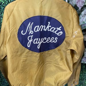 Puede incluir: Chaqueta amarilla mostaza con un parche ovalado azul marino con las palabras "Mankato Jaycees" en escritura blanca. La chaqueta tiene cuello y cierre de botones. La chaqueta está colgada en una percha de madera.