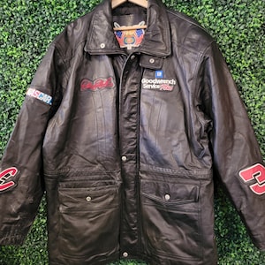May include: Black leather jacket with NASCAR and GM Goodwrench Service Plus logos. Features a collar, multiple pockets, and the number 3 on the sleeve. The jacket is hanging on a wooden hanger.