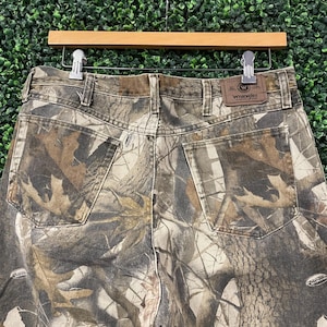 May include: Camouflage-patterned Wrangler jeans, with a brown leather patch bearing the Wrangler logo. The jeans are suspended on a wooden hanger against a green backdrop. The camouflage design incorporates shades of brown, green, and tan.