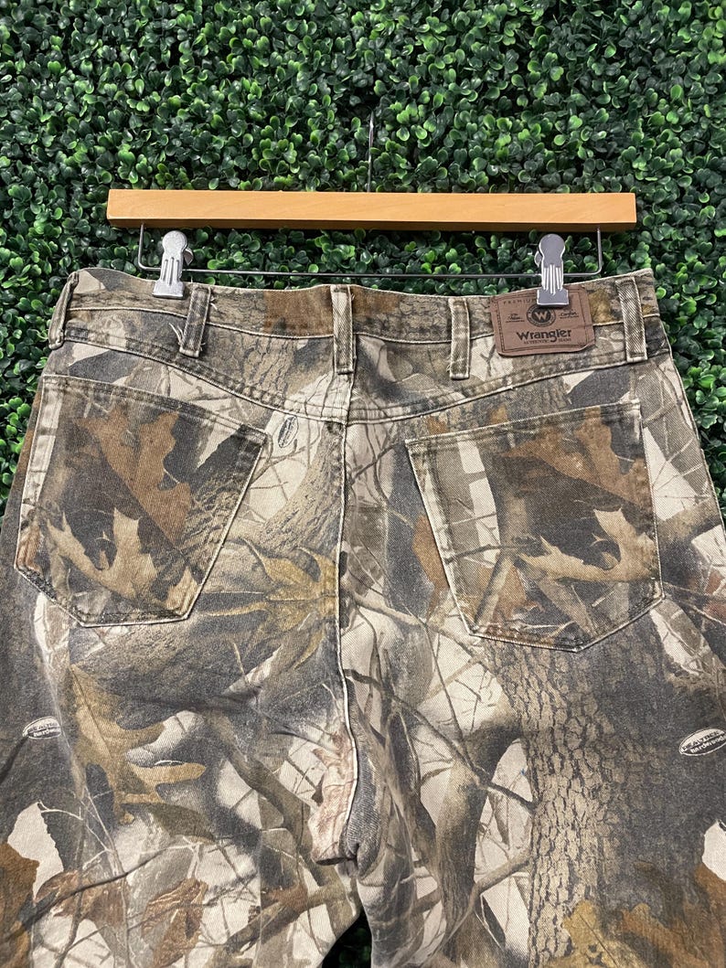 May include: Camouflage-patterned Wrangler jeans, with a brown leather patch bearing the Wrangler logo. The jeans are suspended on a wooden hanger against a green backdrop. The camouflage design incorporates shades of brown, green, and tan.
