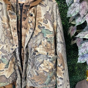 May include: A camouflage jacket with a hood and multiple pockets. The jacket features a brown and green leaf pattern, a black zipper, and snap closures. The jacket is hanging on a wooden hanger.