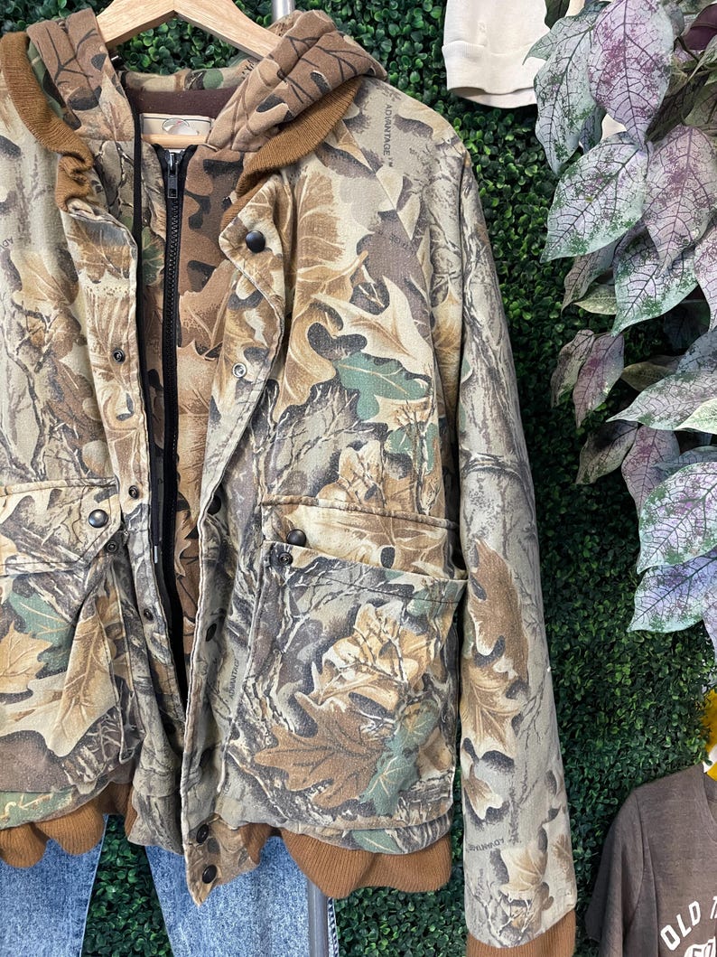 May include: A camouflage jacket with a hood and multiple pockets. The jacket features a brown and green leaf pattern, a black zipper, and snap closures. The jacket is hanging on a wooden hanger.