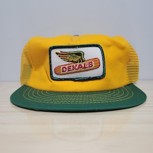 Peut inclure: Casquette de camionneur jaune et verte avec un écusson portant le mot "DEKALB" et un graphisme de maïs ailé. La casquette a un dos en filet et une visière incurvée. La casquette est de style vintage.