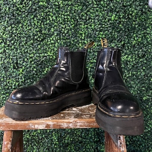 Puede incluir: Par de botas de plataforma de cuero negro con paneles laterales elásticos. Las botas tienen una suela gruesa con costuras amarillas y una lengüeta con texto amarillo. Las botas se exhiben en un pequeño taburete de madera.