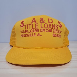 Op de afbeelding: Een gele truckerpet met rode tekst "A & D TITLE LOANS". De pet bevat ook de tekst "CASH LOANS ON CAR TITLES" en de stad "HUNTSVILLE, AL."