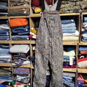 May include: Camouflage-patterned overalls hanging on a wooden hanger. The overalls have adjustable black straps and multiple pockets. The background shows wooden shelves filled with folded clothing, including jeans, sweaters, and jackets.