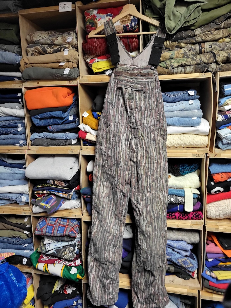 May include: Camouflage-patterned overalls hanging on a wooden hanger. The overalls have adjustable black straps and multiple pockets. The background shows wooden shelves filled with folded clothing, including jeans, sweaters, and jackets.