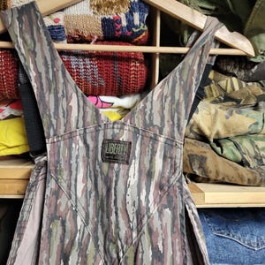 May include: Camouflage-patterned overalls with black straps, hanging on a wooden hanger. The overalls feature a mix of brown, green, and gray vertical stripes. A small label reads "LIBERTY MADE IN USA". The background shows shelves with folded clothing.