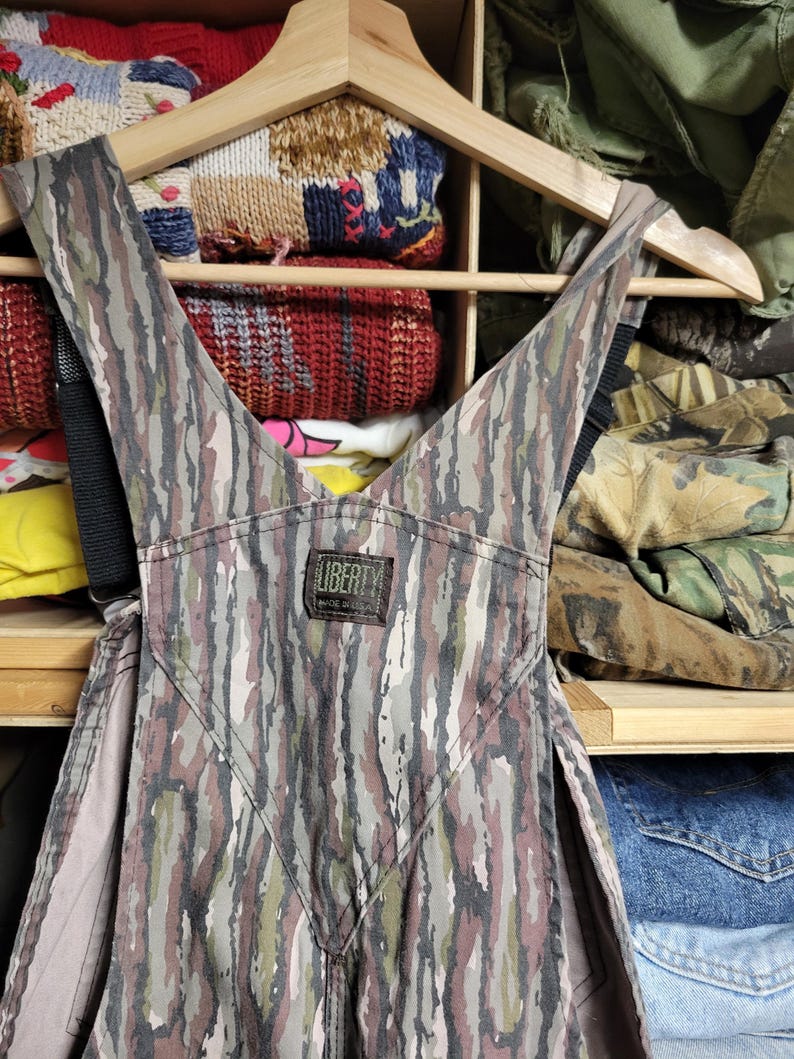 May include: Camouflage-patterned overalls with black straps, hanging on a wooden hanger. The overalls feature a mix of brown, green, and gray vertical stripes. A small label reads "LIBERTY MADE IN USA". The background shows shelves with folded clothing.