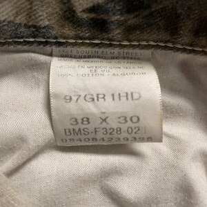 May include: A close-up of a label from camouflage-patterned jeans. The label indicates the size as 96.5 x 76 cm, along with the style number and other details. The jeans are made of 100% cotton.