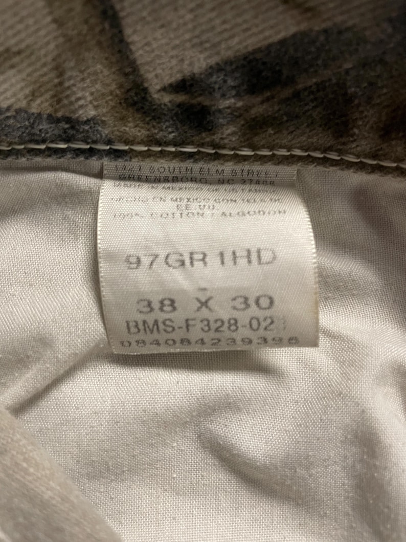 May include: A close-up of a label from camouflage-patterned jeans. The label indicates the size as 96.5 x 76 cm, along with the style number and other details. The jeans are made of 100% cotton.