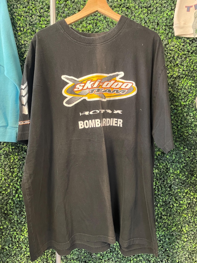 May include: Black t-shirt with a graphic that reads "Ski-Doo Team" in yellow, orange, and white. Below the logo are the words "Rotax" and "Bombardier" in white. The shirt is hanging on a wooden hanger.