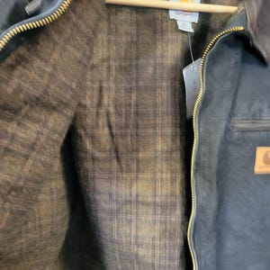 May include: A dark navy blue jacket with a brass-colored metal zipper and a brown plaid flannel lining. The interior features a white Carhartt brand label at the neck. A tan rectangular leather patch with the Carhartt logo is visible on the front.
