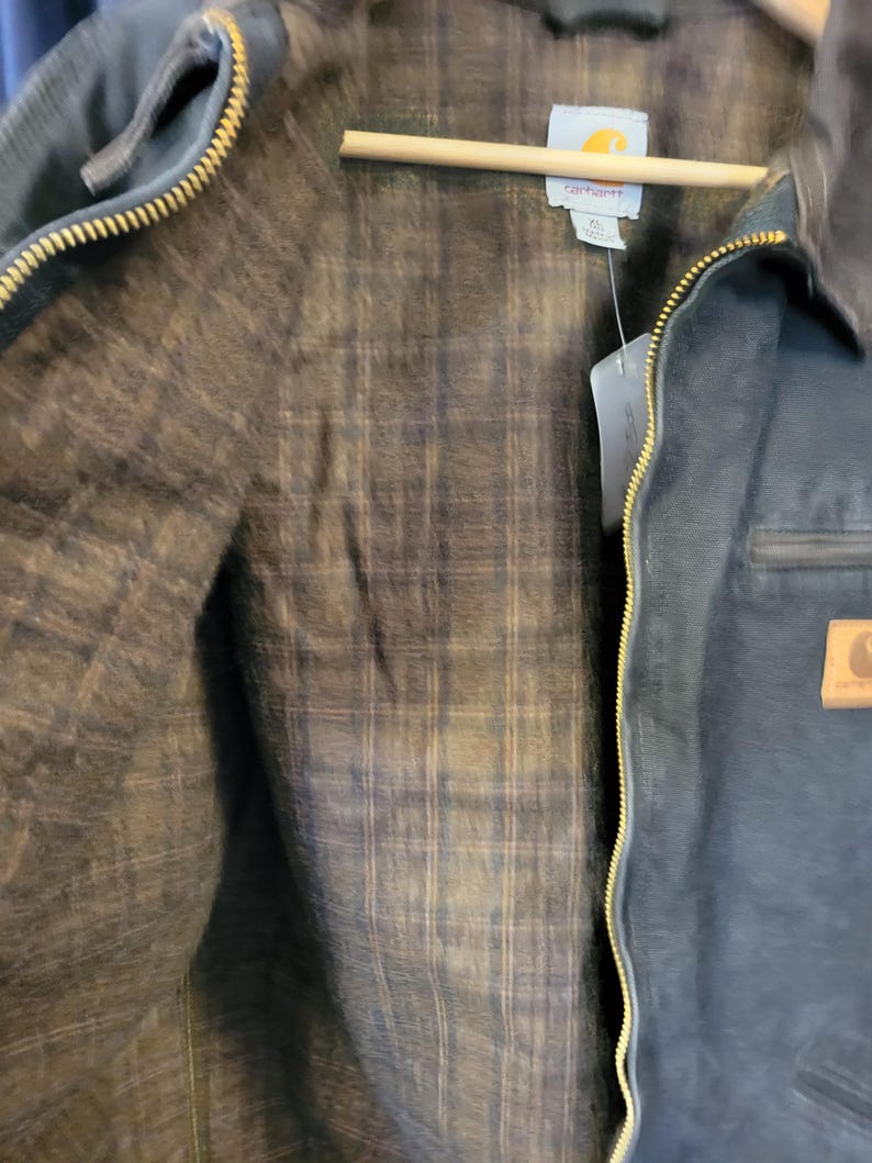 May include: A dark navy blue jacket with a brass-colored metal zipper and a brown plaid flannel lining. The interior features a white Carhartt brand label at the neck. A tan rectangular leather patch with the Carhartt logo is visible on the front.