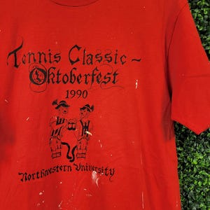 May include: Red t-shirt with black text that reads "Tennis Classic - Oktoberfest 1990" and "Northwestern University." The shirt features a graphic of two figures in traditional German attire. The shirt has white paint splatters.