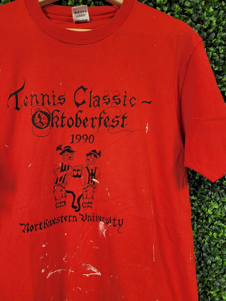 May include: Red t-shirt with black text that reads "Tennis Classic - Oktoberfest 1990" and "Northwestern University." The shirt features a graphic of two figures in traditional German attire. The shirt has white paint splatters.