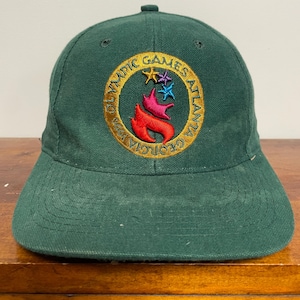 Peut inclure: Une casquette de baseball vert foncé avec un logo circulaire brodé indiquant Olympic Games Atlanta 1996 Georgia. Le centre de l'emblème présente un motif de flamme stylisé en rouge, rose et bleu, surmonté de deux petites étoiles.