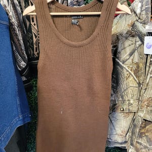 Puede incluir: Un top sin mangas marrón acanalado colgado de una percha de madera. El top sin mangas tiene cuello redondo y una silueta ajustada. La tela parece ser de punto. El top sin mangas es talla L.