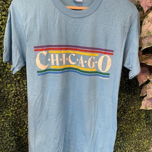 Peut inclure: T-shirt bleu clair avec le mot "CHICAGO" en blanc, souligné de couleurs arc-en-ciel. Le t-shirt est suspendu à un cintre en bois. Col rond et manches courtes.