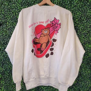 May include: A white long-sleeve sweatshirt featuring a graphic of a brown teddy bear sitting inside a red heart-shaped box with a pink bow. The text "How do I Love THEE..." is printed above the heart, surrounded by small brown chocolate candies.