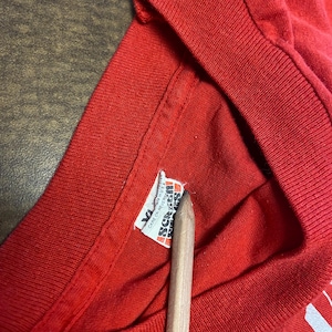 Puede incluir: Primer plano de un cuello de camiseta roja con una etiqueta. La etiqueta dice "XL" y "Screen Stars" con instrucciones de cuidado. Un l&aacute;piz de madera apunta a la etiqueta.