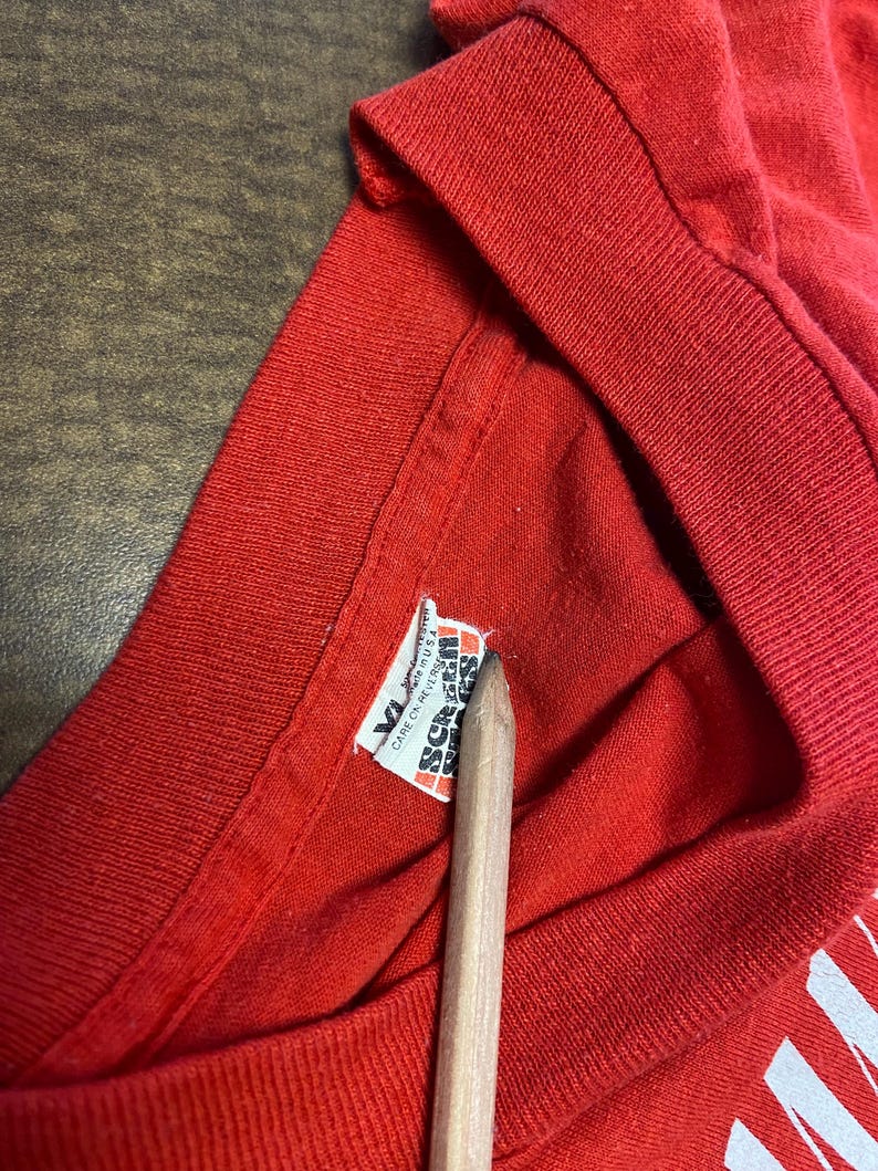 Puede incluir: Primer plano de un cuello de camiseta roja con una etiqueta. La etiqueta dice "XL" y "Screen Stars" con instrucciones de cuidado. Un l&aacute;piz de madera apunta a la etiqueta.