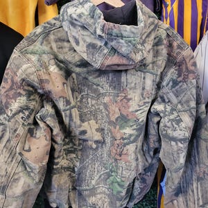 May include: A camouflage hooded jacket with a black interior lining and a black ribbed hem. The jacket features a woodland pattern with shades of brown, green, and tan. The jacket is hanging on a wooden hanger.