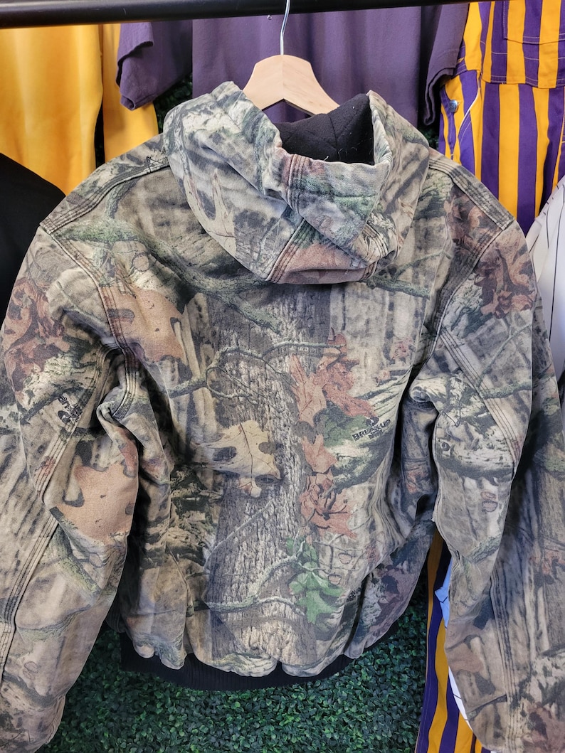 May include: A camouflage hooded jacket with a black interior lining and a black ribbed hem. The jacket features a woodland pattern with shades of brown, green, and tan. The jacket is hanging on a wooden hanger.