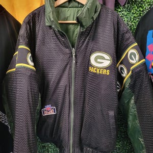Puede incluir: Chaqueta reversible de los Packers en negro y verde con cremallera completa. La chaqueta presenta el logotipo de los Packers en el pecho y los colores del equipo en las mangas y el cuello. El parche de la NFL Experience es visible en la parte delantera.