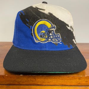 Könnte beinhalten: Eine Baseballkappe mit einer blau-weißen Krone und einem schwarzen Pinselstrich-Design. Auf der Vorderseite befindet sich ein gesticktes Logo eines gelb-blauen Footballhelms. Die Kappe hat einen schwarzen flachen Schirm und eine grüne Unterseite.