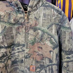 May include: A camouflage hooded jacket with a full zipper closure. The jacket features a brown Carhartt logo patch on the front pocket and a distressed appearance. The pattern includes shades of brown, green, and tan, resembling a forest environment.