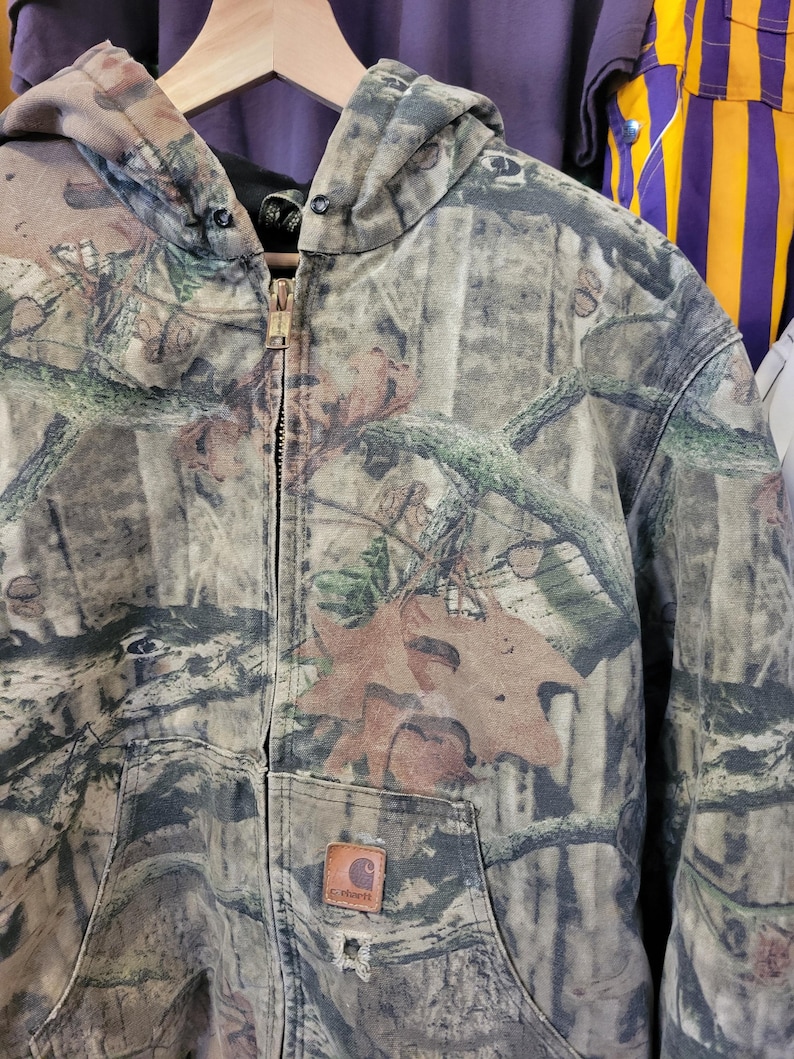 May include: A camouflage hooded jacket with a full zipper closure. The jacket features a brown Carhartt logo patch on the front pocket and a distressed appearance. The pattern includes shades of brown, green, and tan, resembling a forest environment.