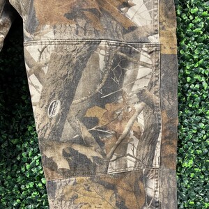 May include: Close-up of camouflage trousers with a detailed pattern of brown, tan, and grey tree branches and foliage. The trousers have reinforced pockets and stitching, with a small logo patch visible. The fabric appears durable and suitable for outdoor wear.