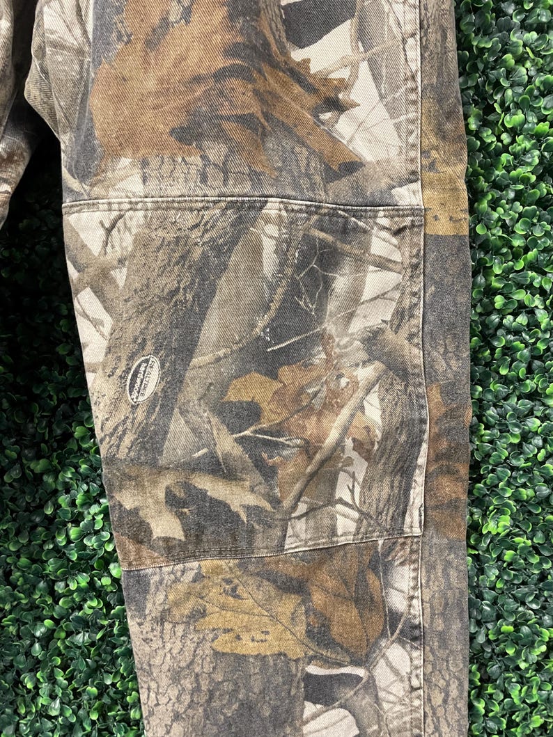 May include: Close-up of camouflage trousers with a detailed pattern of brown, tan, and grey tree branches and foliage. The trousers have reinforced pockets and stitching, with a small logo patch visible. The fabric appears durable and suitable for outdoor wear.