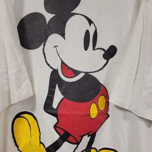 K&ouml;nnte beinhalten: Wei&szlig;es T-Shirt mit einer gro&szlig;en Mickey Mouse-Grafik. Mickey wird in seiner klassischen Pose gezeigt, mit schwarzen Ohren, einem wei&szlig;en Gesicht, roten Shorts, gelben Schuhen und einem l&auml;chelnden Gesichtsausdruck. Das T-Shirt hat kurze &Auml;rmel.