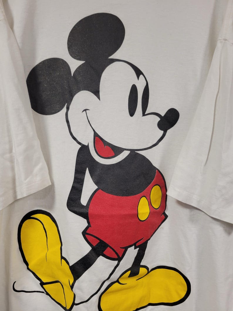 K&ouml;nnte beinhalten: Wei&szlig;es T-Shirt mit einer gro&szlig;en Mickey Mouse-Grafik. Mickey wird in seiner klassischen Pose gezeigt, mit schwarzen Ohren, einem wei&szlig;en Gesicht, roten Shorts, gelben Schuhen und einem l&auml;chelnden Gesichtsausdruck. Das T-Shirt hat kurze &Auml;rmel.