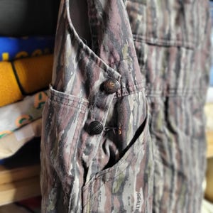 May include: Close-up of a camouflage overall with a buttoned pocket. The fabric features a pattern of brown, green, and gray. The overall has a pocket with a button closure and the word "Realtree" is visible.