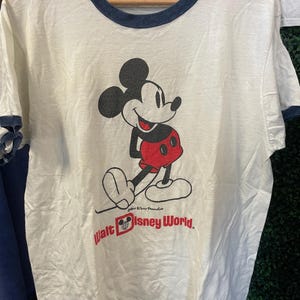May include: A white ringer t-shirt with dark blue trim on the collar and sleeves. The front features a graphic of Mickey Mouse in black and red, with the text Walt Disney World printed in red below the character, including a small Mickey icon.