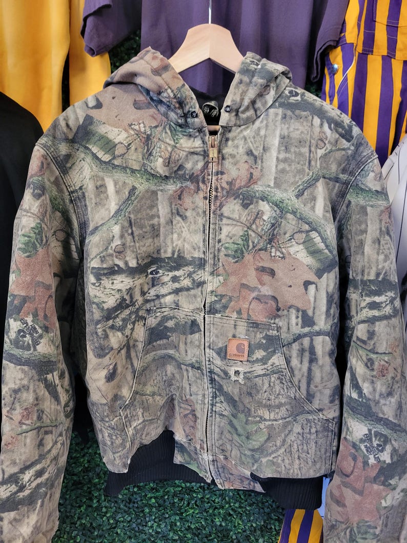 May include: A camouflage hooded jacket with a full zipper closure and two front pockets. The jacket features a brown, green, and tan pattern, and a Carhartt logo patch. The jacket is hanging on a wooden hanger.