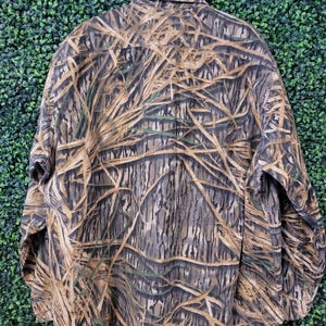 May include: A long-sleeved button-down shirt with a camouflage pattern in shades of brown, tan, and green, resembling reeds and branches. The shirt has a classic collar and buttoned cuffs, suitable for outdoor activities.