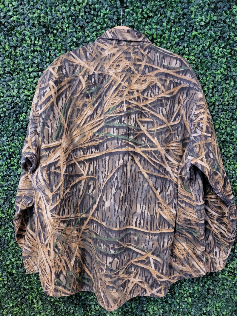 May include: A long-sleeved button-down shirt with a camouflage pattern in shades of brown, tan, and green, resembling reeds and branches. The shirt has a classic collar and buttoned cuffs, suitable for outdoor activities.