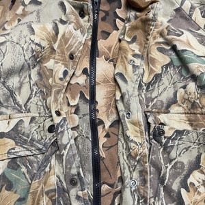 May include: A camouflage jacket with a full-length black zipper and a brown hood. The jacket features a pattern of leaves in shades of brown, green, and tan. It has multiple pockets and button closures.