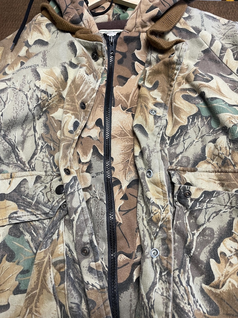 May include: A camouflage jacket with a full-length black zipper and a brown hood. The jacket features a pattern of leaves in shades of brown, green, and tan. It has multiple pockets and button closures.