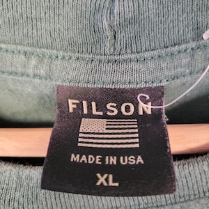 May include: Close-up of a green t-shirt with a black tag. The tag reads "FILSON" above an American flag graphic and "MADE IN USA" with "XL" below.